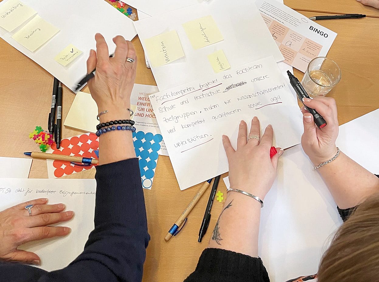 Zwei Personen lehnen über einen Tisch mit vielen Zetteln, Klebepunkten etc. und schreiben gerade an einem kurzen Text.